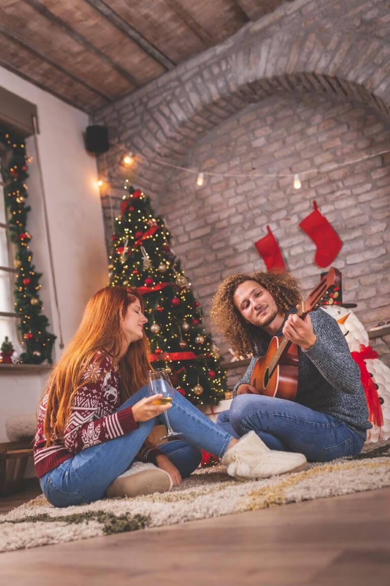 Zwei Personen sitzen auf dem Boden in einem Raum. Eine Person spielt Gitarre und der Raum ist Weihnachtlich geschmückt
